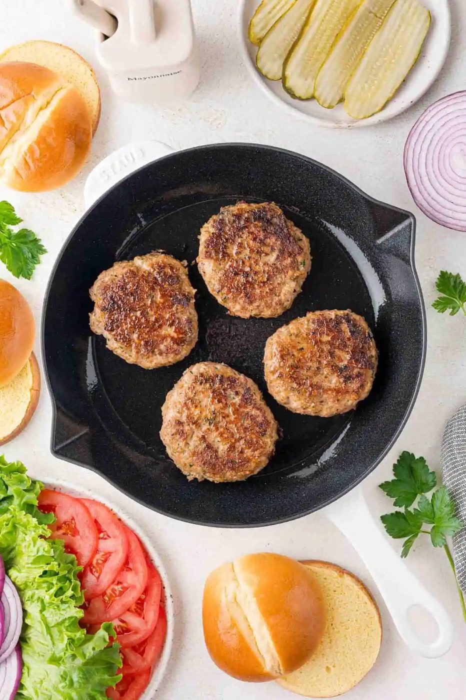 Four cooked turkey burger patties in a white-handled skillet surrounded by burger buns, lettuce, tomato slices, pickles, and red onion slices on a light surface.
