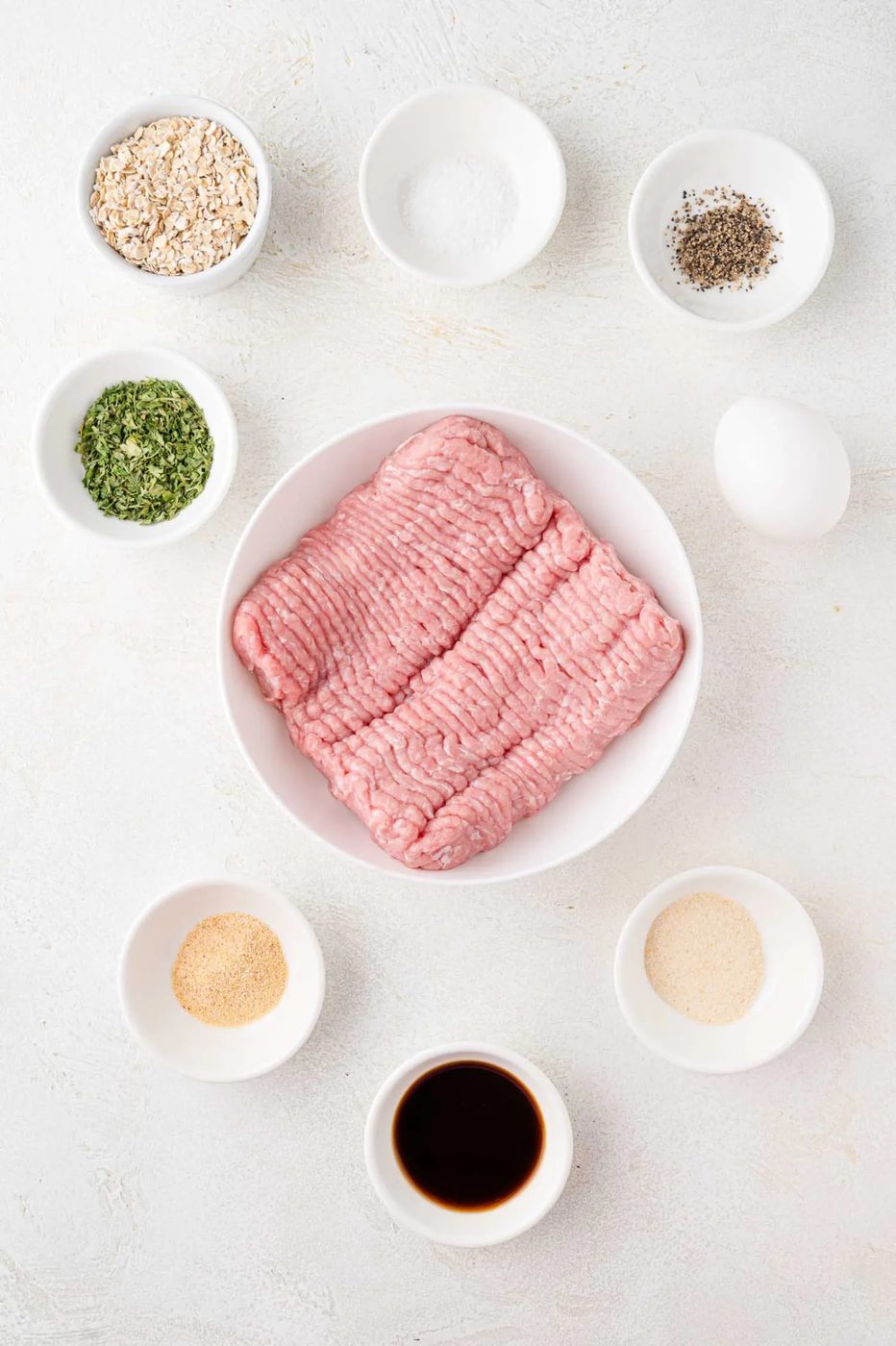 A bowl of ground meat surrounded by small bowls containing oats, salt, pepper, and other ingredients on a light surface.