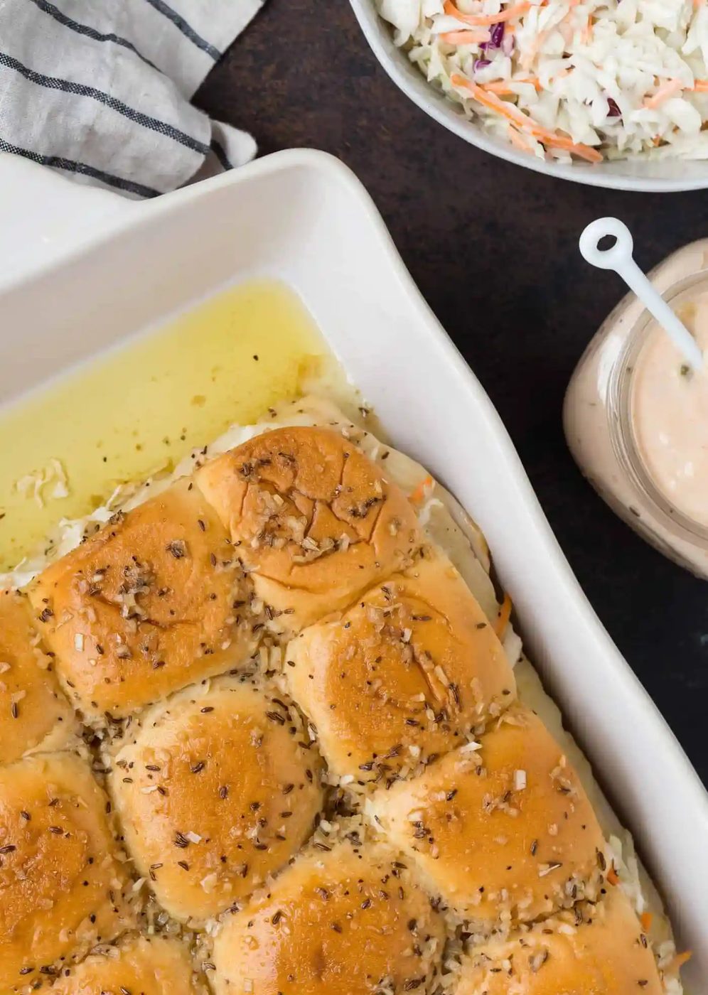 Overhead view of butter-soaked slider buns in a white baking dish. They are filled with coleslaw, turkey, swiss cheese, and thousand island dressing.