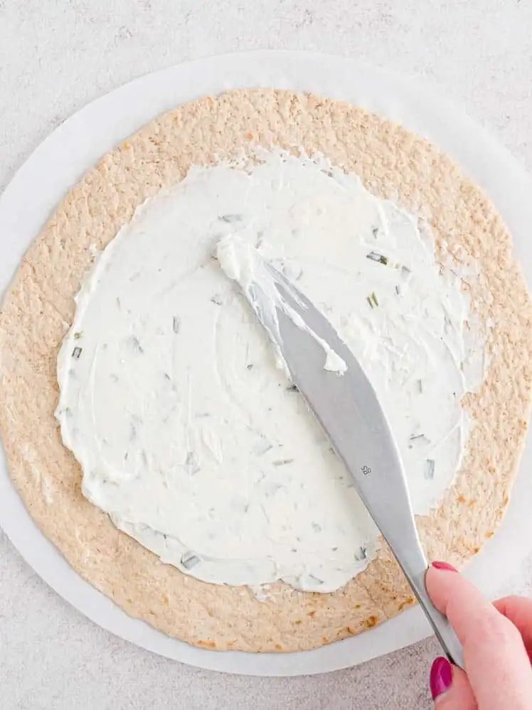 A hand using a knife to spread onion and chive cream cheese over a flour tortilla.