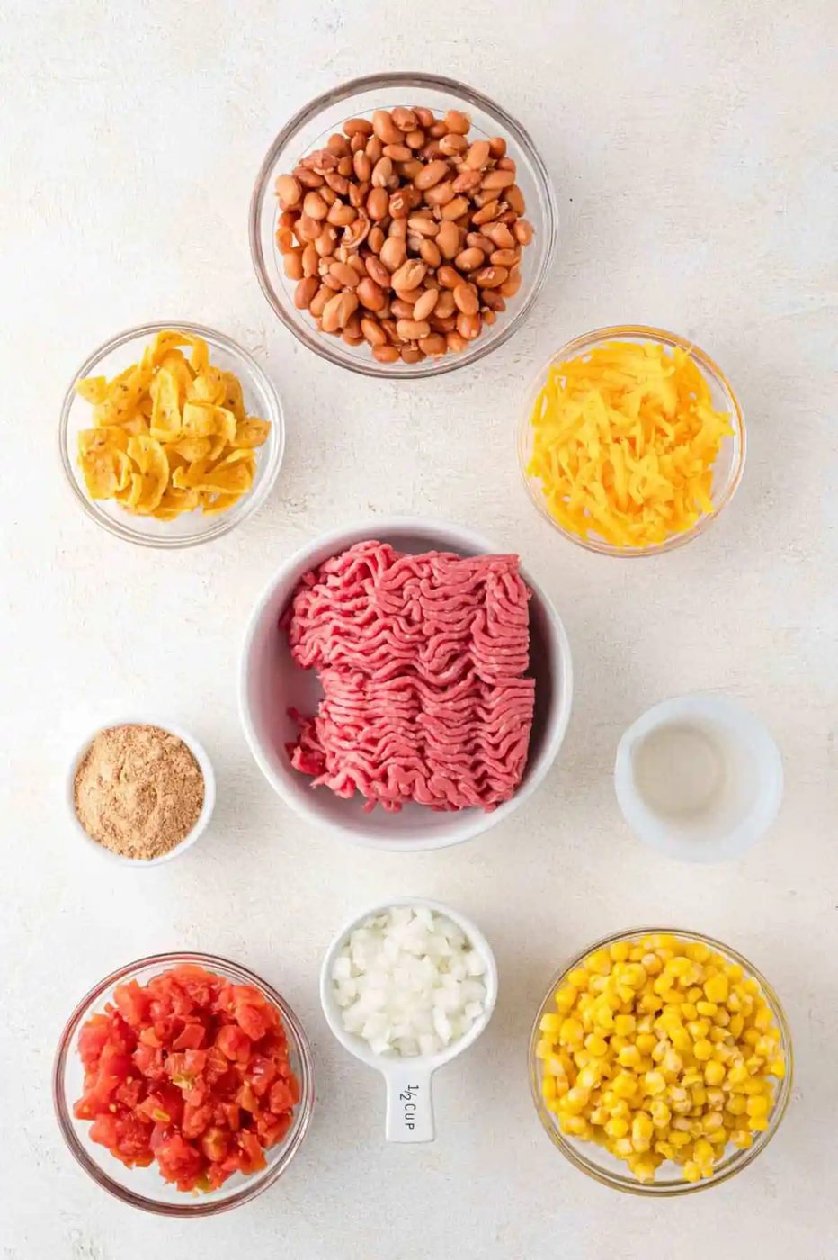 Bowls of ingredients arranged on a surface, including raw ground beef, beans, shredded cheese, corn, chopped tomatoes, crushed chips, onions, a spice mix, and a small dish of water.