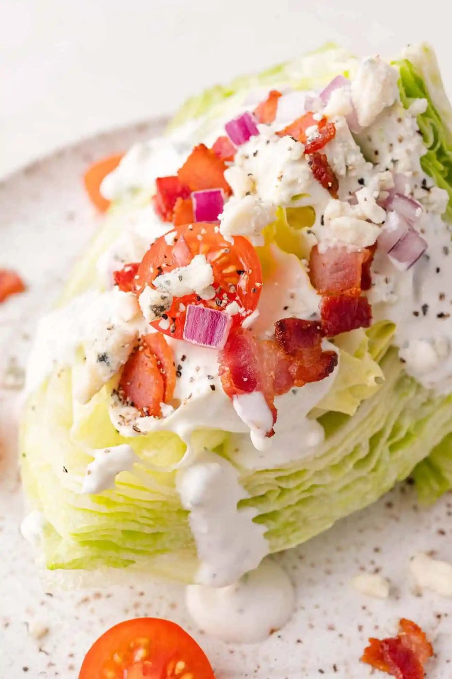 A wedge salad topped with cherry tomatoes, red onion, bacon, and crumbled cheese, drizzled with a creamy dressing on a speckled plate.