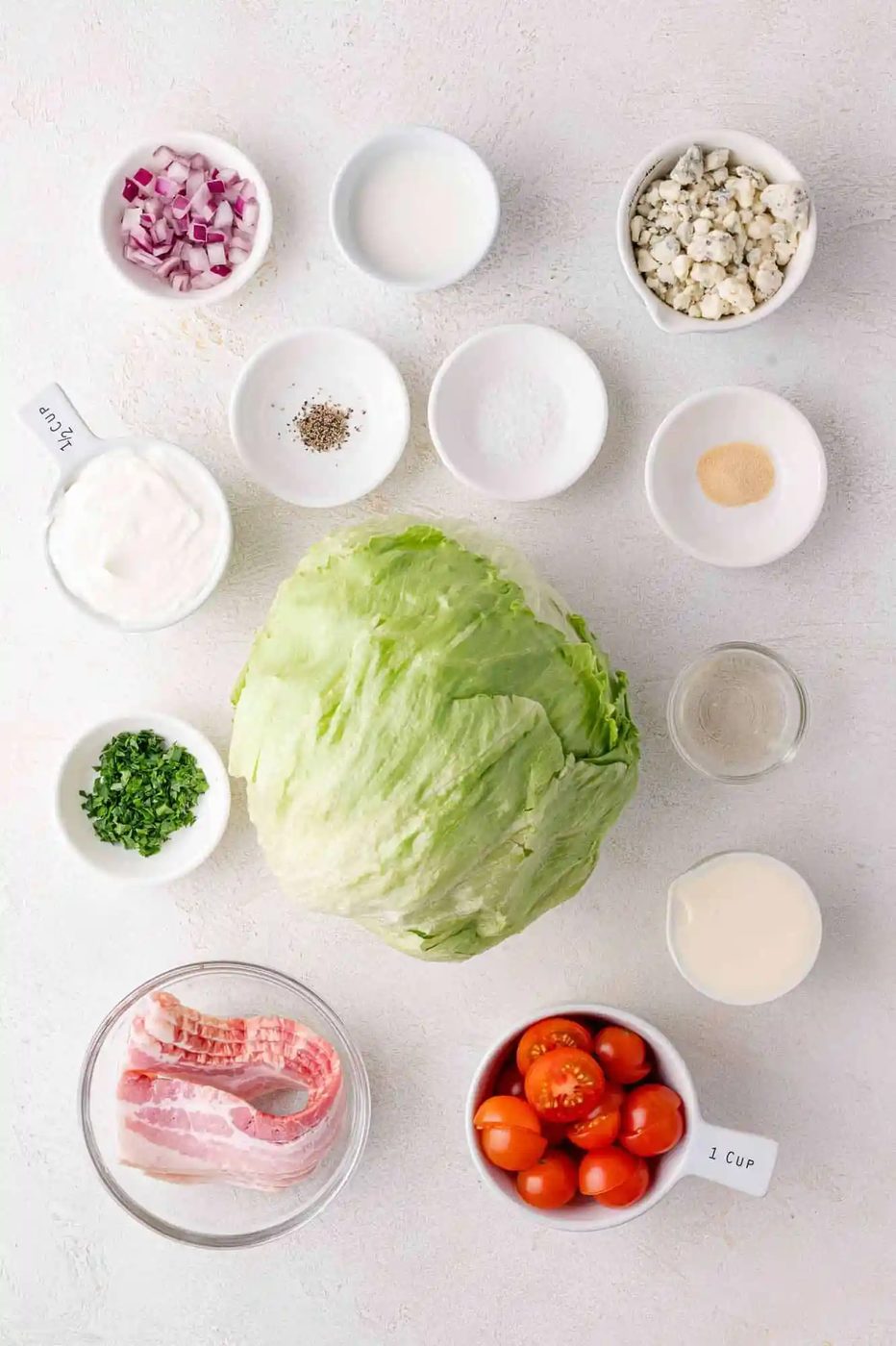 Ingredients for a salad include iceberg lettuce, cherry tomatoes, bacon, red onion, blue cheese, sour cream, parsley, and assorted seasonings, arranged on a white surface.