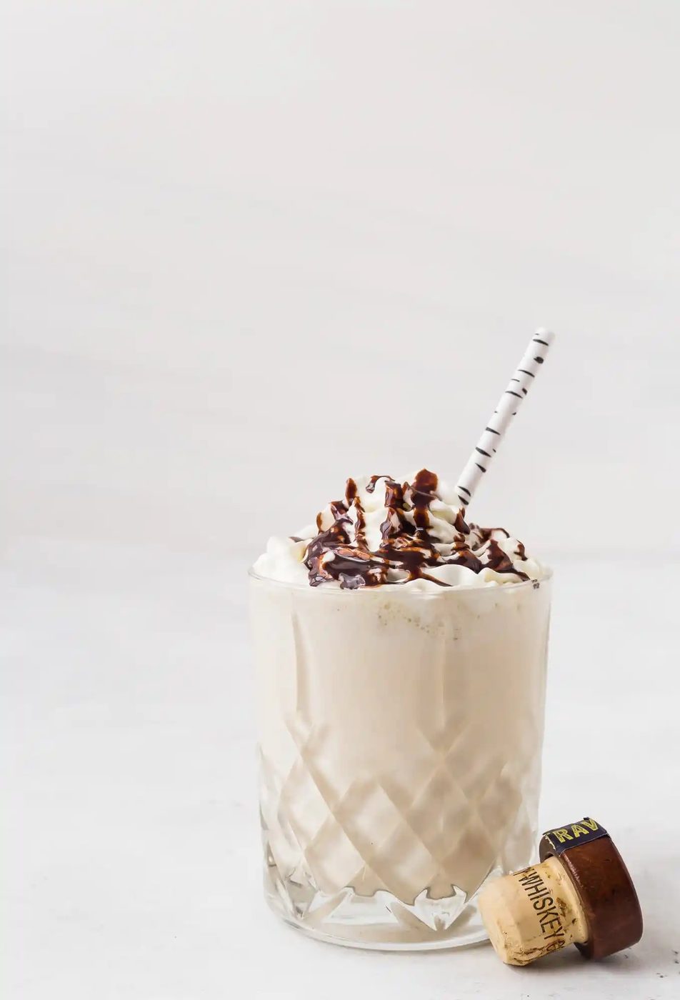 A glass of creamy milkshake topped with whipped cream and chocolate drizzle, with a striped straw and a bottle cap beside the glass.
