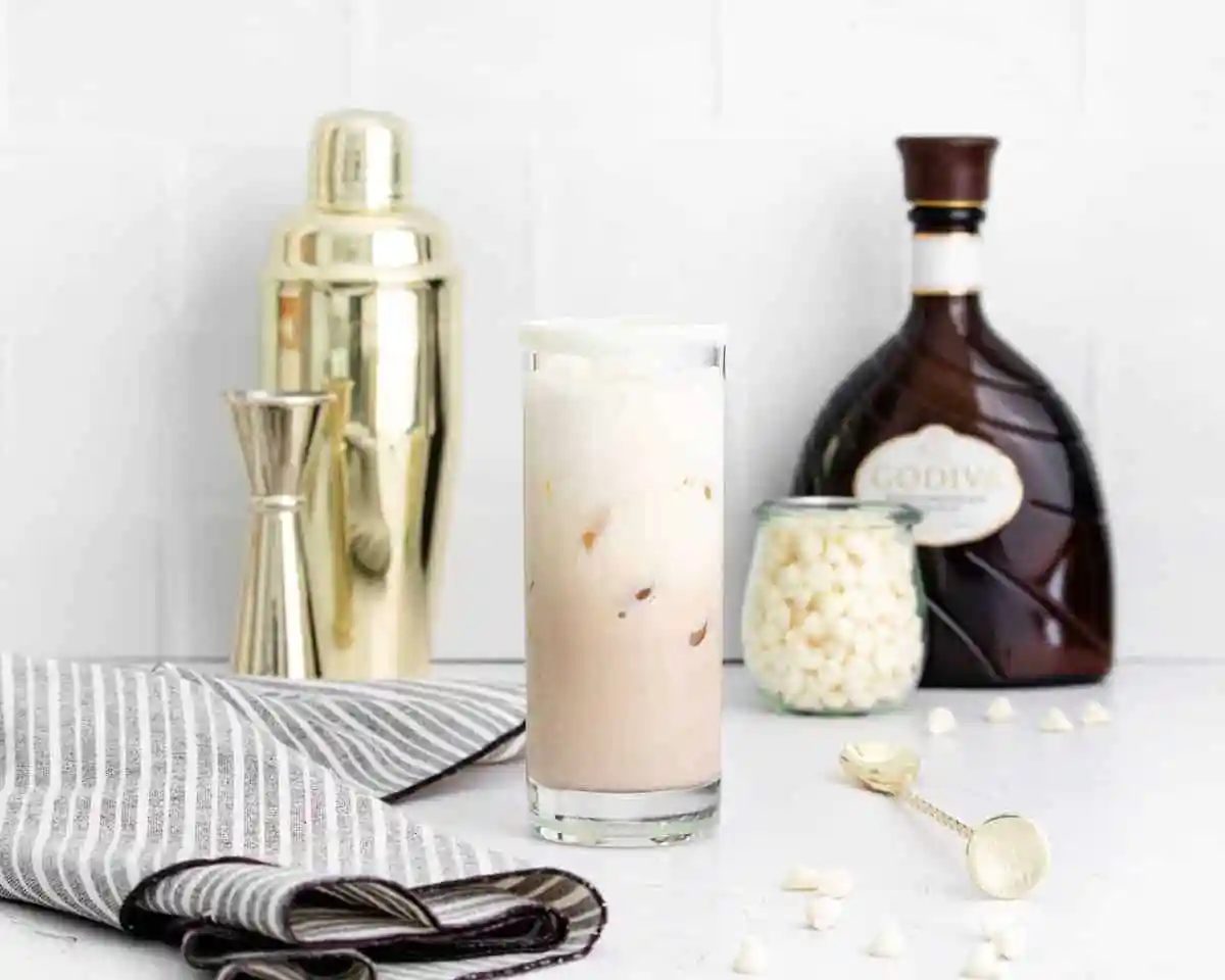 A cocktail in a glass with ingredients and a shaker in the background.