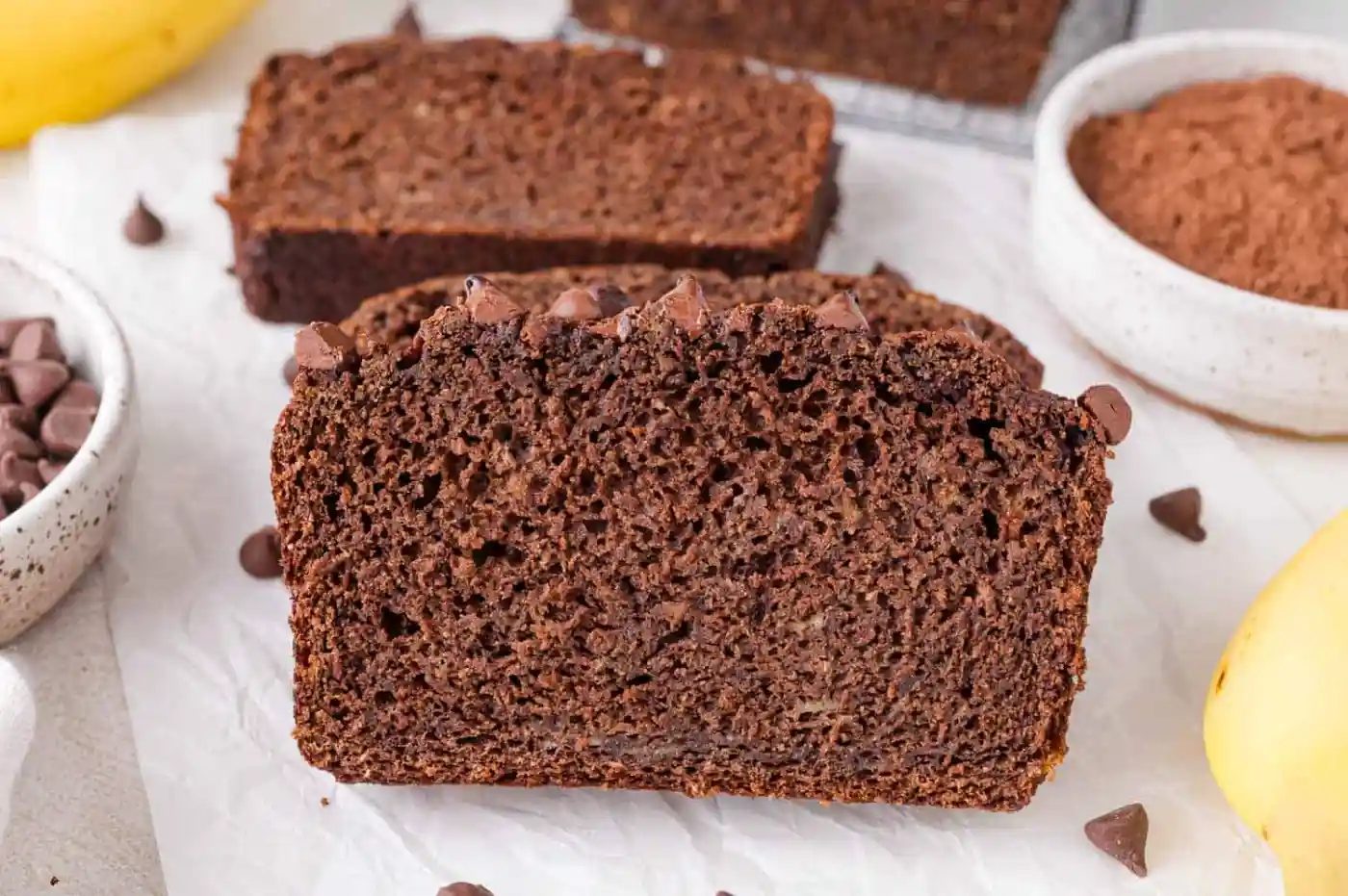 A close-up of a sliced loaf of lighter chocolate banana bread, with chocolate chips visible, surrounded by a bowl of cocoa powder, chocolate chips, and bananas.