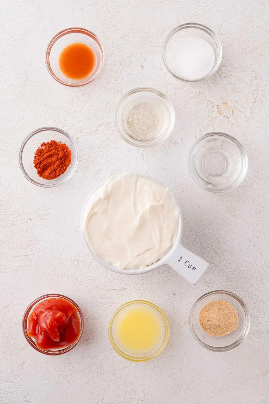 Nine small bowls containing various ingredients, including mayonnaise, ketchup, hot sauce, spices, and liquids, arranged on a white surface.