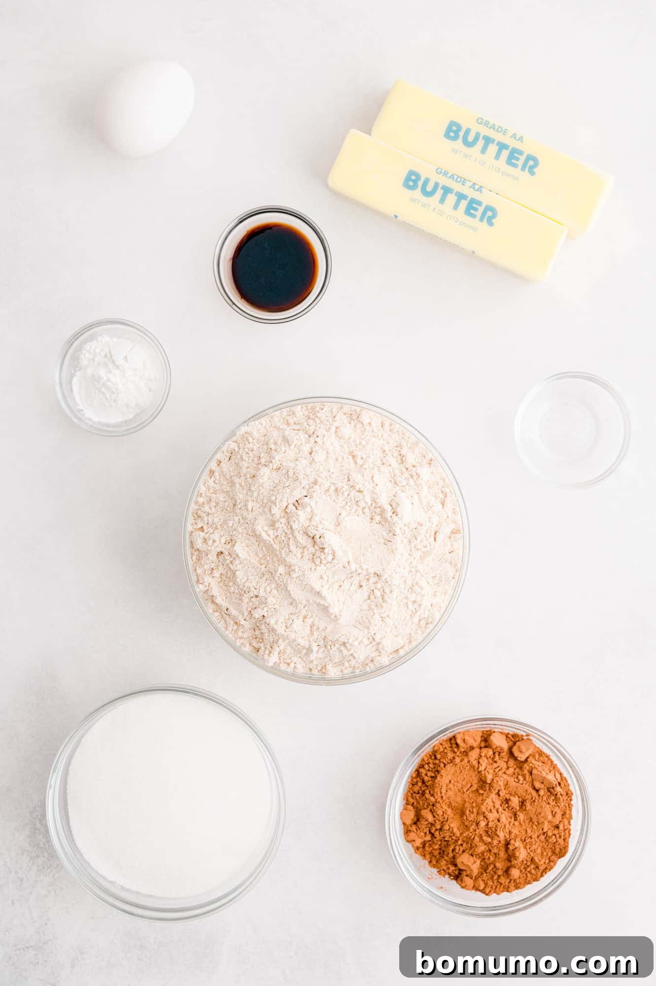 All the essential ingredients for peppermint chocolate sugar cookies, including butter, sugar, flour, and cocoa powder, neatly arranged in separate bowls on a pristine white background, ready for baking.