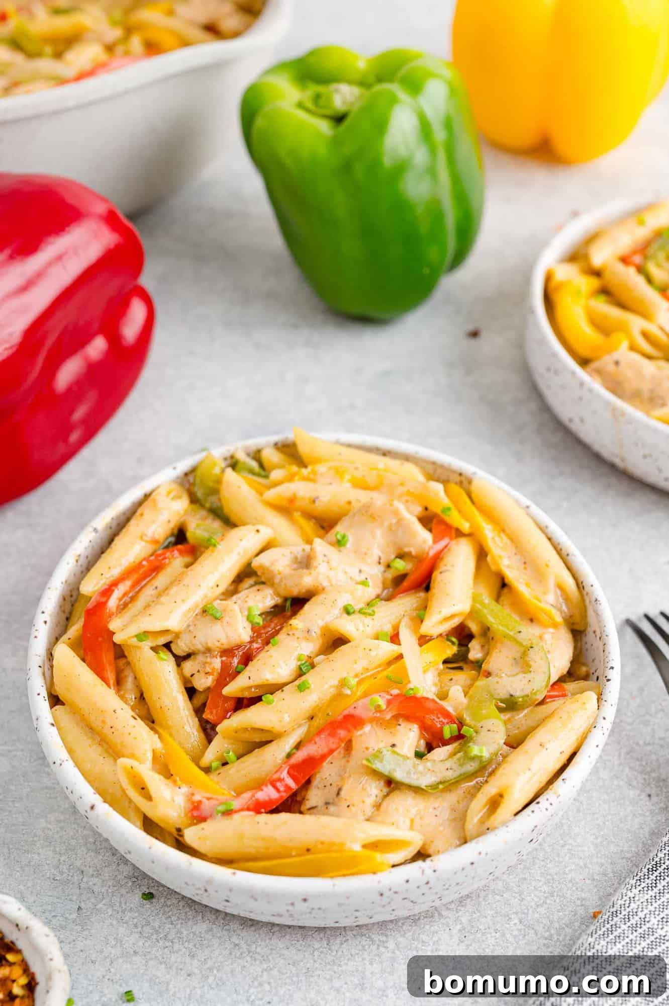 A bowl of Rasta Pasta next to red, green, and yellow bell peppers on a countertop.