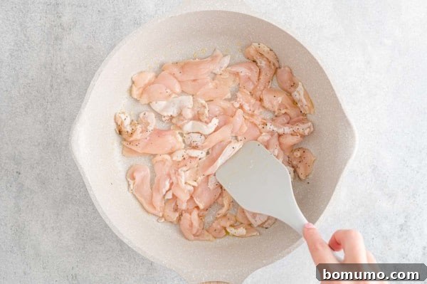 A hand uses a spatula to stir chicken pieces in a skillet.