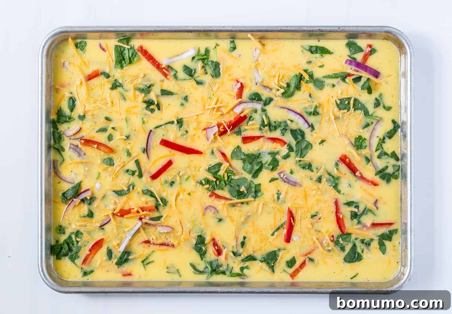 Egg mixture in a sheet pan, generously topped with a colorful assortment of vegetables and shredded cheese, ready for baking.