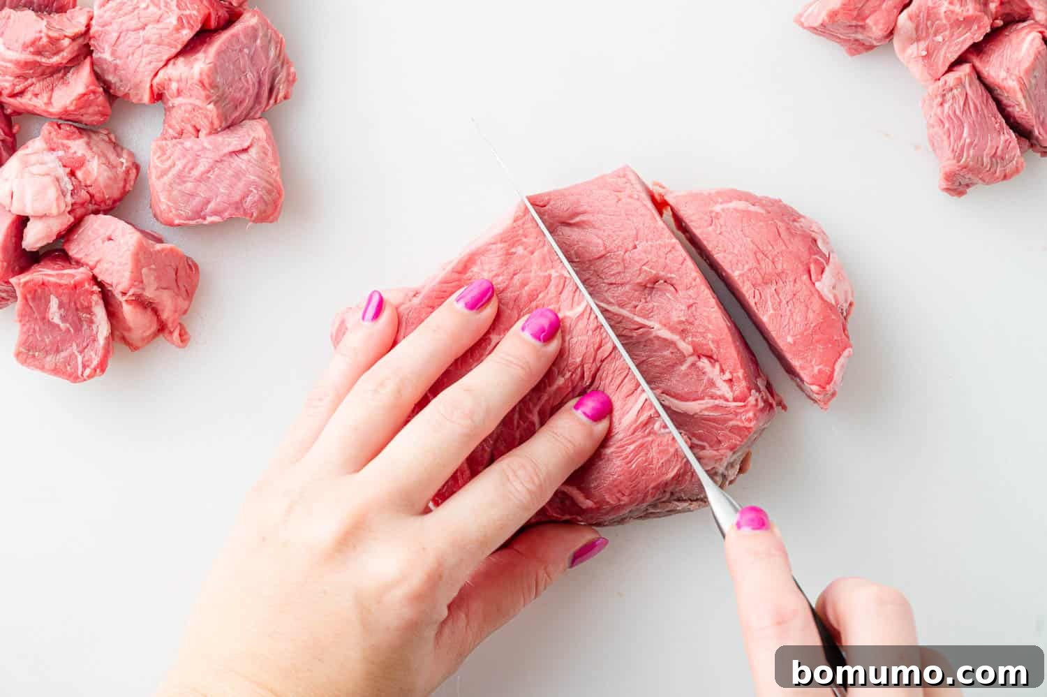 Raw sirloin steak being precisely cut into uniform bite-sized pieces on a cutting board.