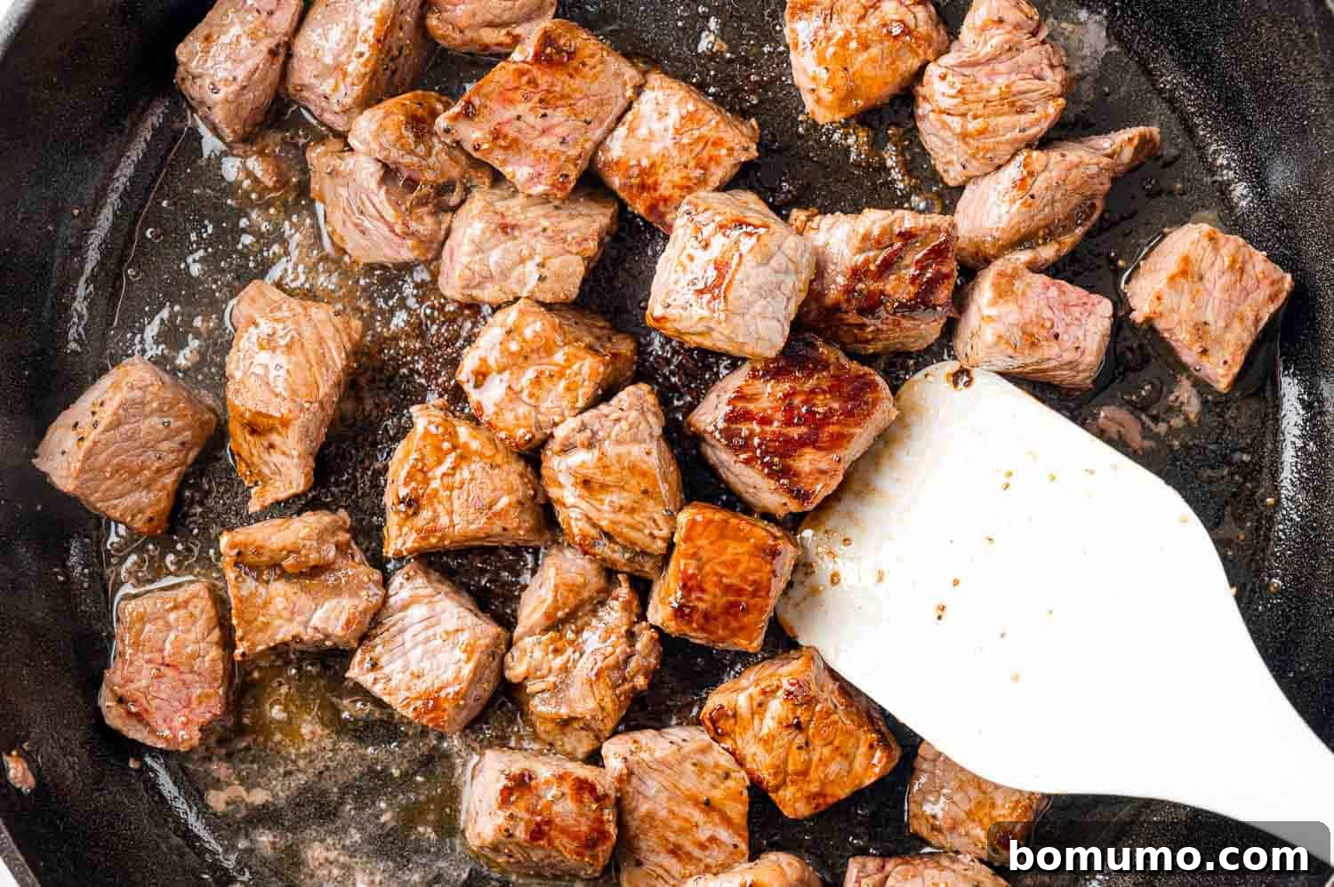 Fully cooked steak bites, perfectly browned and tender, being stirred in the pan with a white spatula.