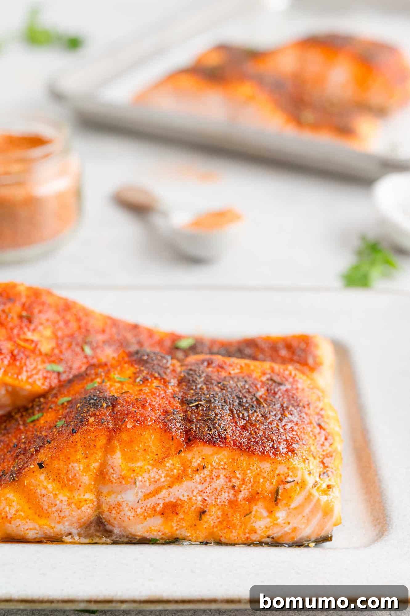 Close up side view of broiled salmon filets on a plate, with more filets on a foil-lined baking sheet in the background.
