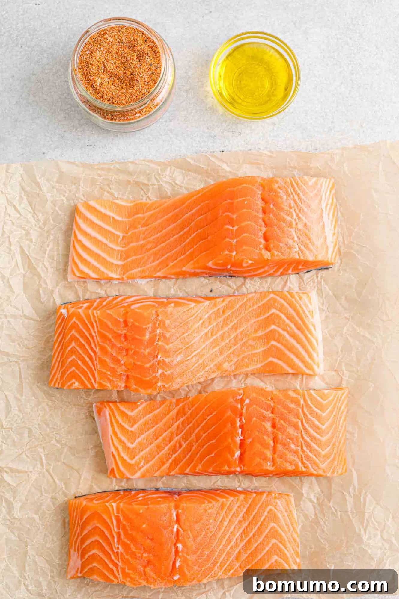 Four salmon filets on a sheet of parchment paper next to small bowls of olive oil and salmon seasoning.