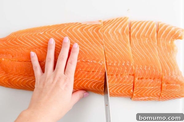A hand holds a side of salmon in place while the other hand uses a sharp knife to cut the salmon into filets.