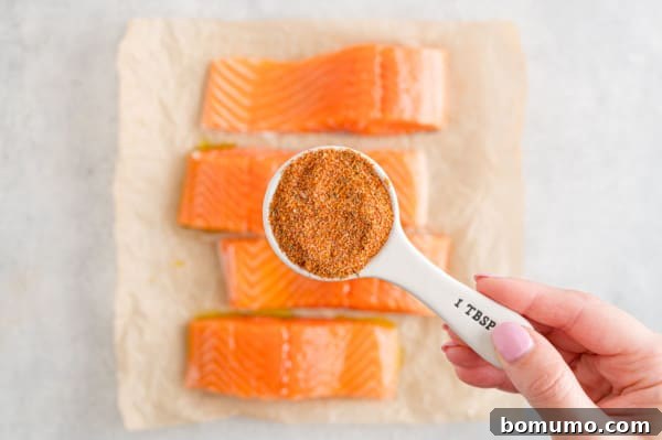 A hand holding a tablespoon of salmon seasoning over four salmon filets rubbed with olive oil.