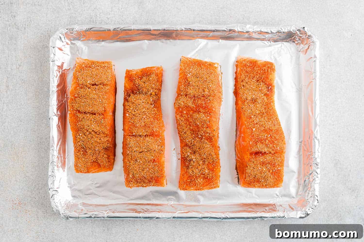 Four seasoned salmon filets on a foil-lined baking sheet.