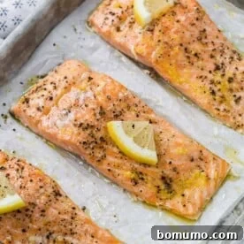 Three slow roasted salmon fillets topped with bright lemon slices and cracked black pepper on a parchment-lined baking sheet.