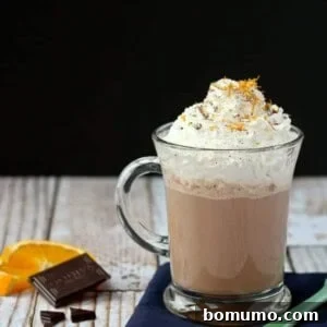Front view of hot chocolate in a clear glass mug, garnished with whipped cream, and fresh orange zest, showing its rich texture.