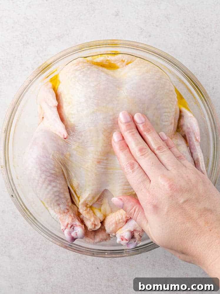 A hand rubbing oil onto a whole raw chicken in a glass bowl on a light-colored surface.