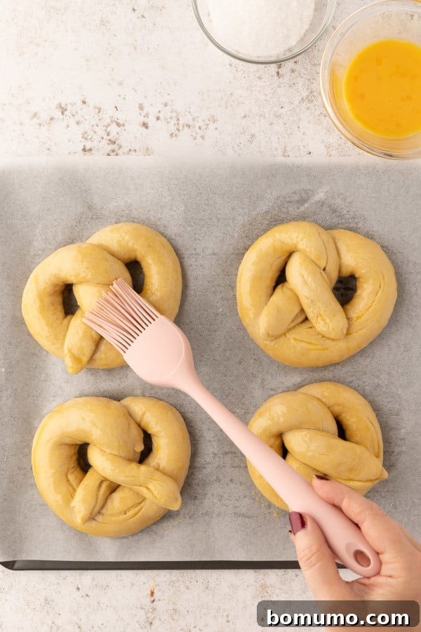 Pretzels on a baking sheet being brushed with an egg wash for a shiny, golden finish.
