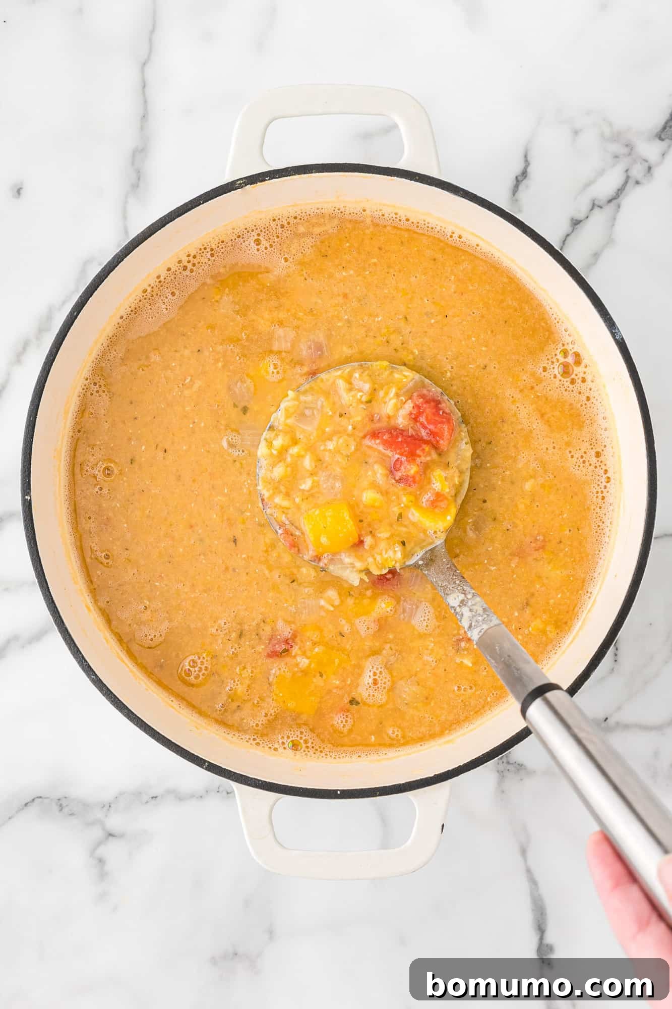 A close-up view of a ladle showing the creamy consistency of the red lentil soup, with visible chunks of vegetables.