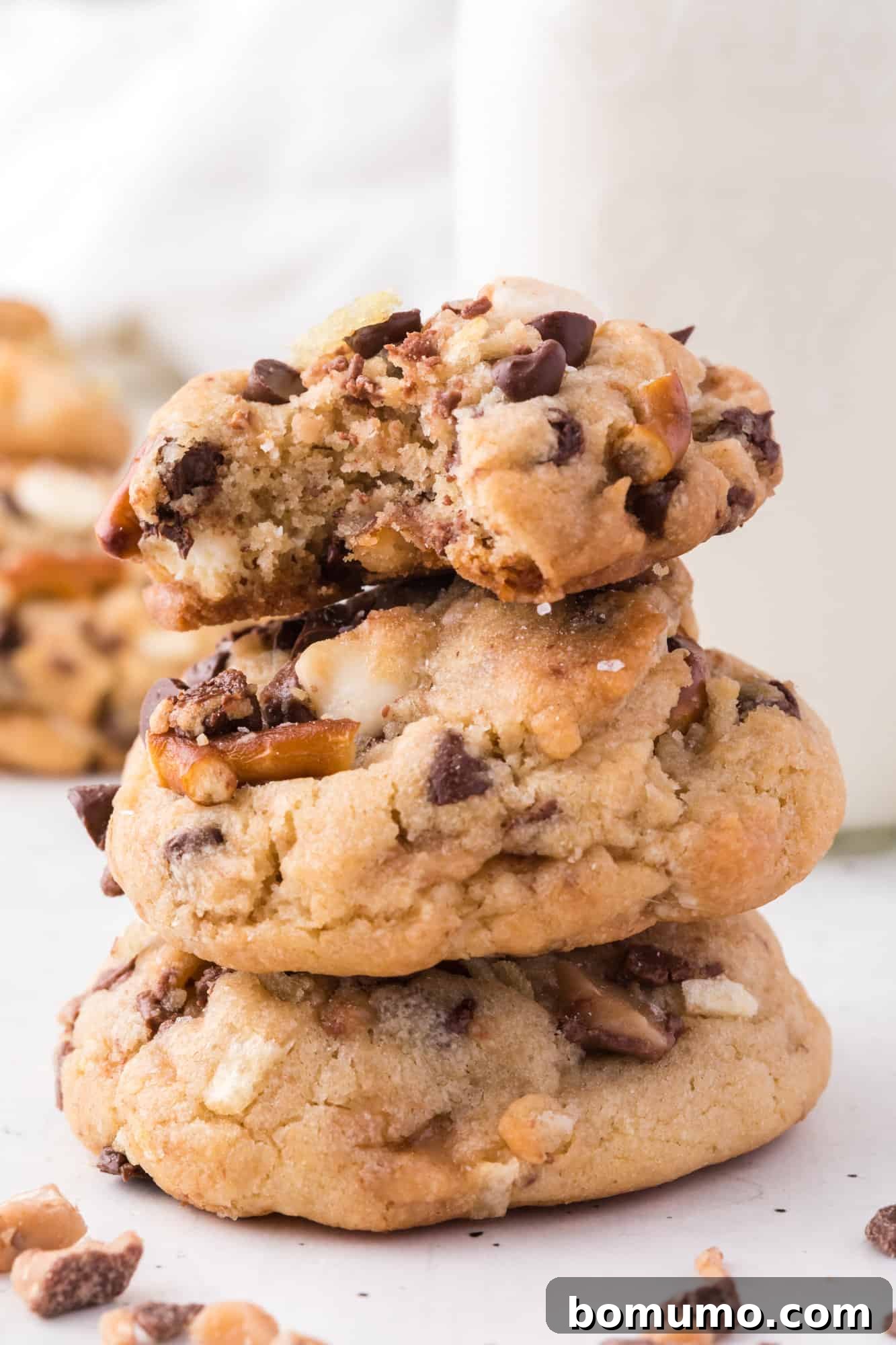 A perfect stack of three golden-brown kitchen sink cookies artfully arranged on a countertop, with a blurred glass of milk in the background.