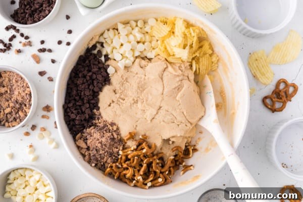 Mixed add-ins, including chocolate chips, pretzels, potato chips, and toffee, folded into a large bowl of vanilla cookie dough.