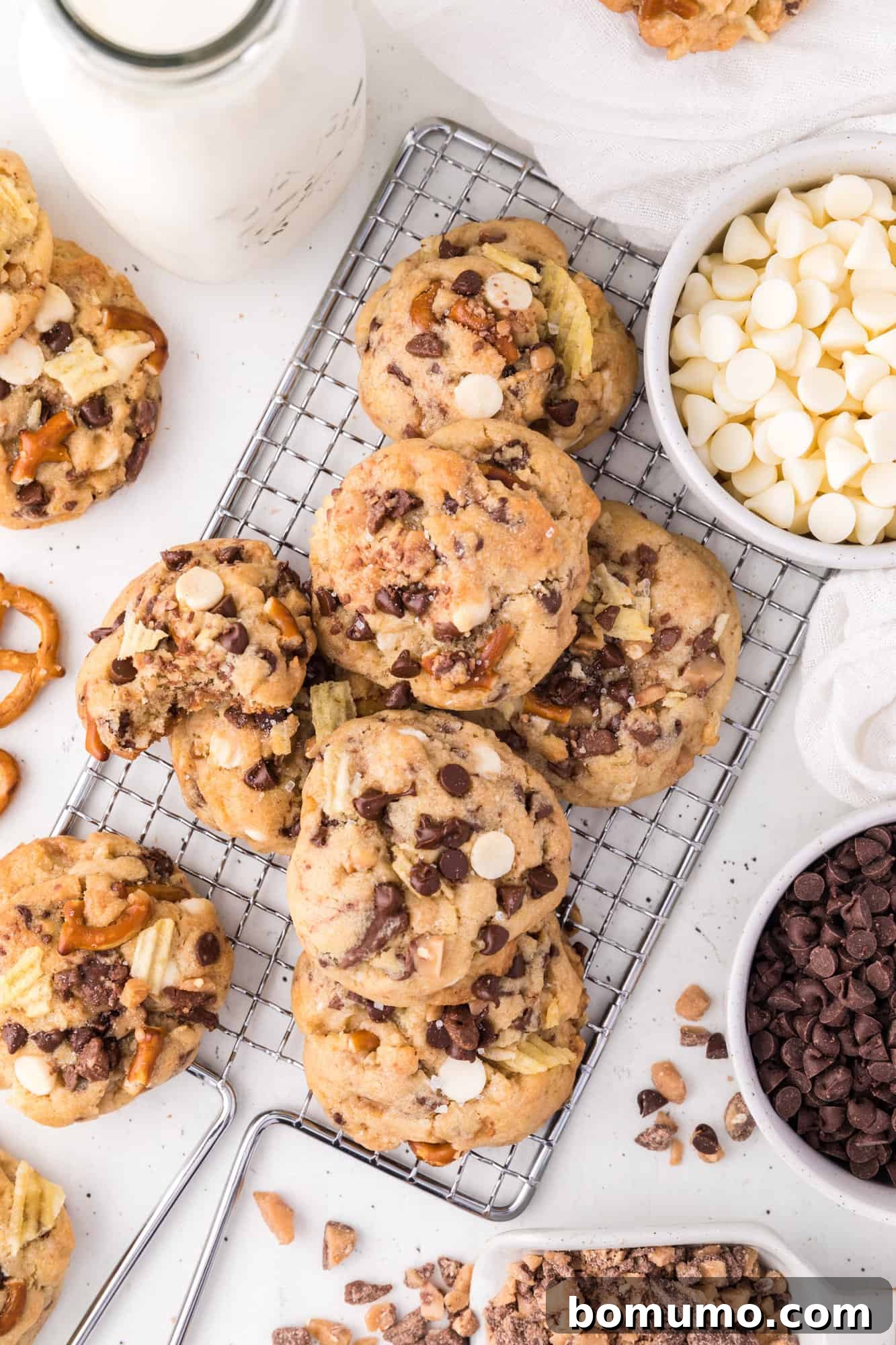 An inviting assortment of freshly baked kitchen sink cookies piled artfully on a wire rack, surrounded by small bowls of their delicious add-ins.