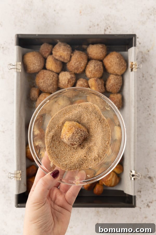 A hand holding a bowl of cinnamon sugar with a partially coated pretzel bite, over more pretzel bites on a baking sheet.