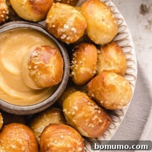 Overhead view of a pretzel bite dipped in a bowl of cheese sauce, surrounded by more pretzel bites on a white plate.