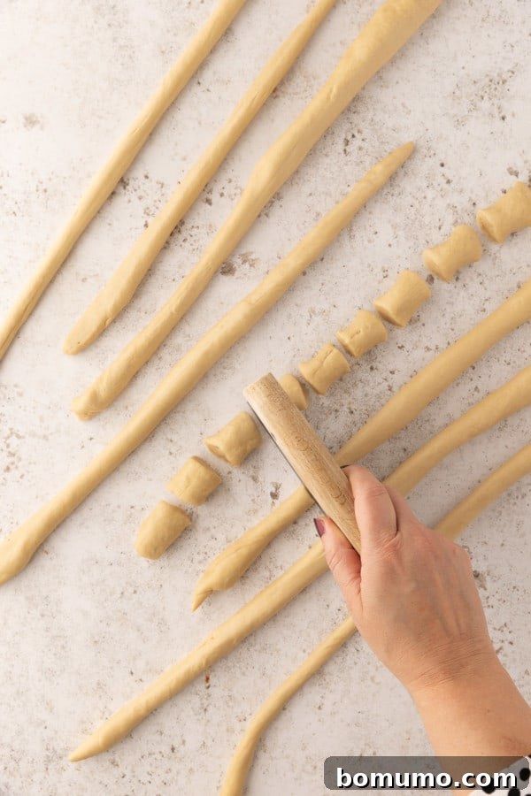 A hand using a dough cutter to cut ropes of pretzel dough into small bite-sized pieces.