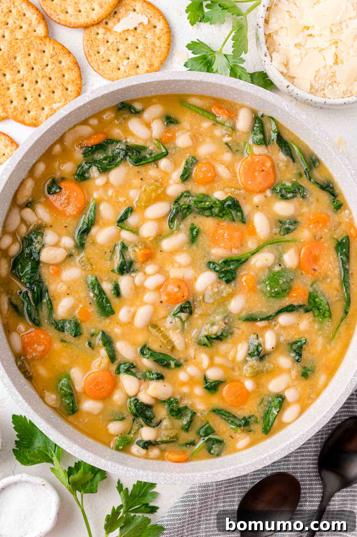 A pot of soup with white beans, carrots, and spinach. Crackers, parsley, and two spoons are nearby.
