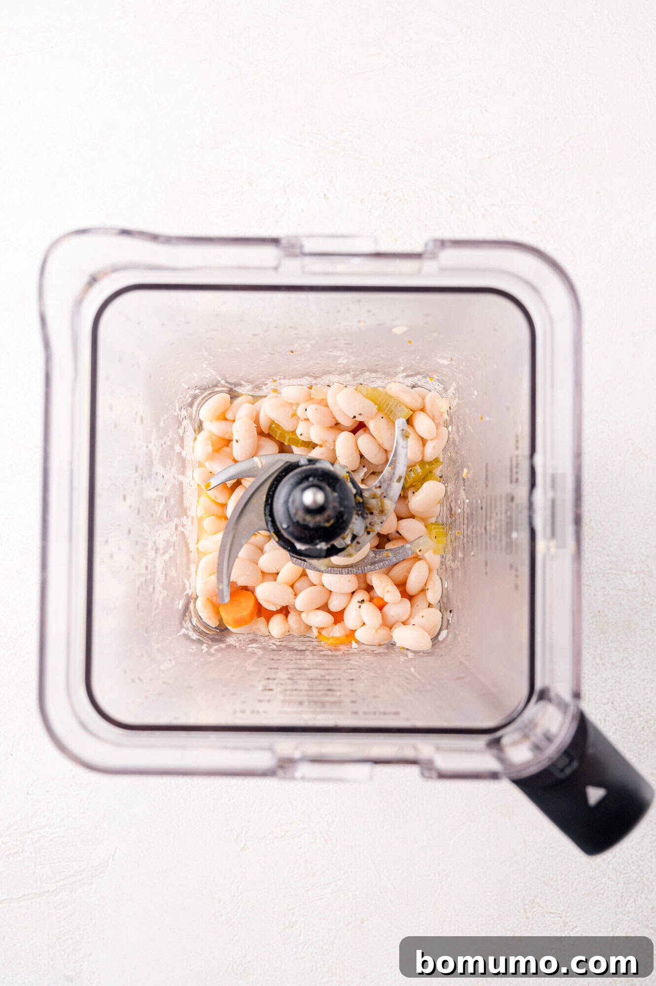 Top-down view of a blender with white beans and sliced carrots inside, ready for blending.