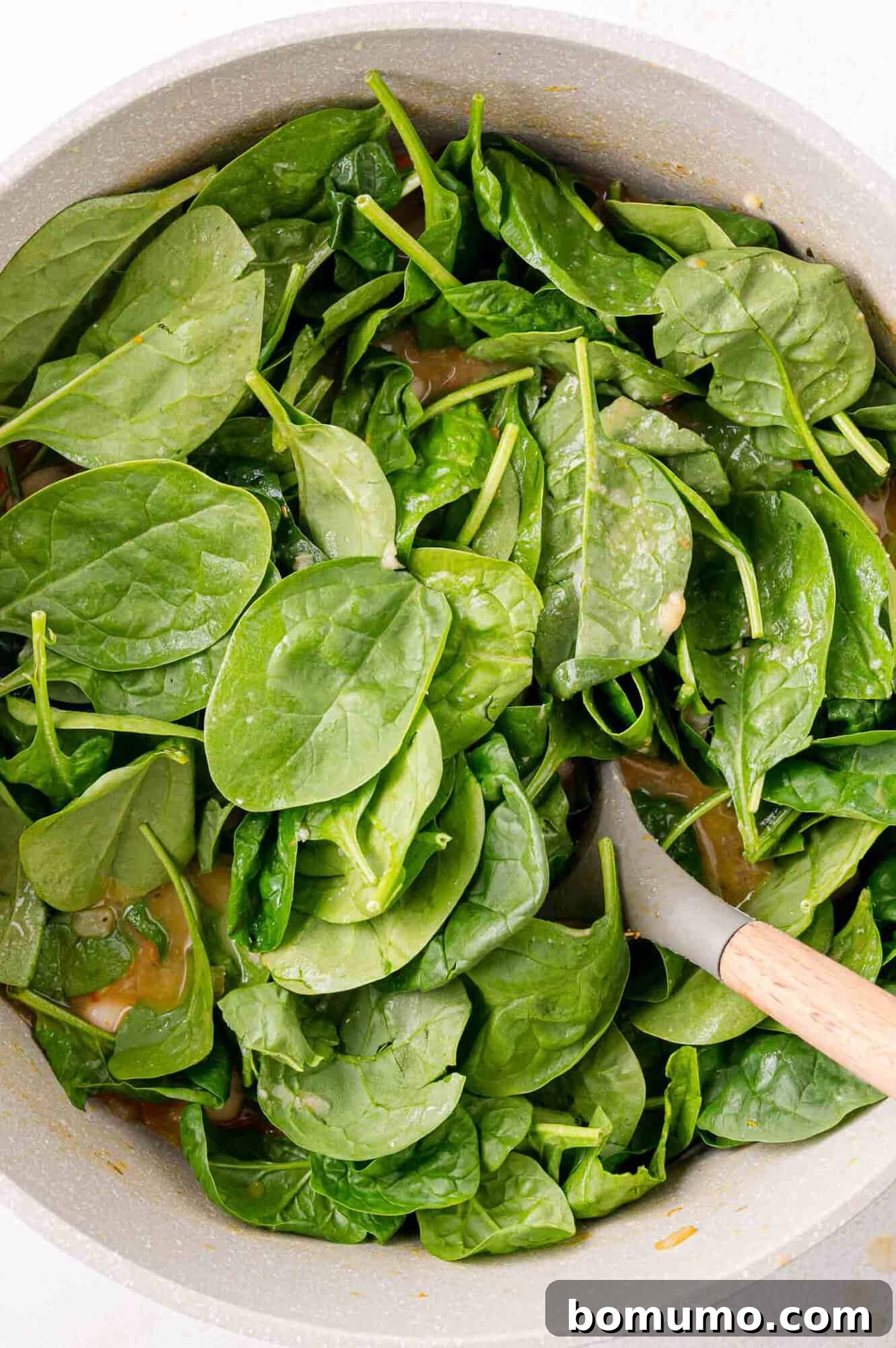 A pan filled with fresh spinach leaves being sautéed, with a wooden spoon resting inside.