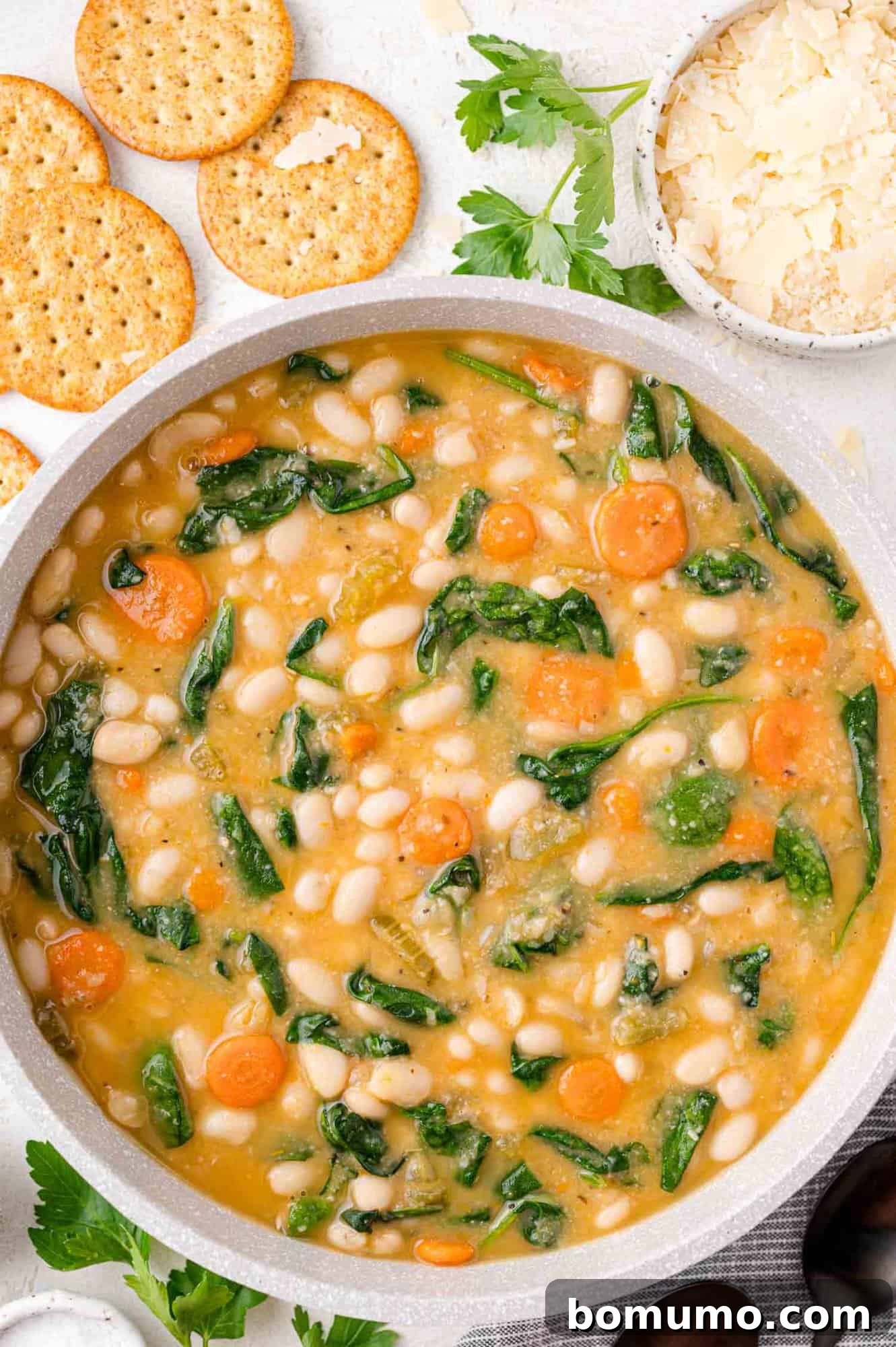 A bowl of white bean and vegetable soup with carrots and spinach, garnished with parsley. Crackers and grated cheese are on the side.