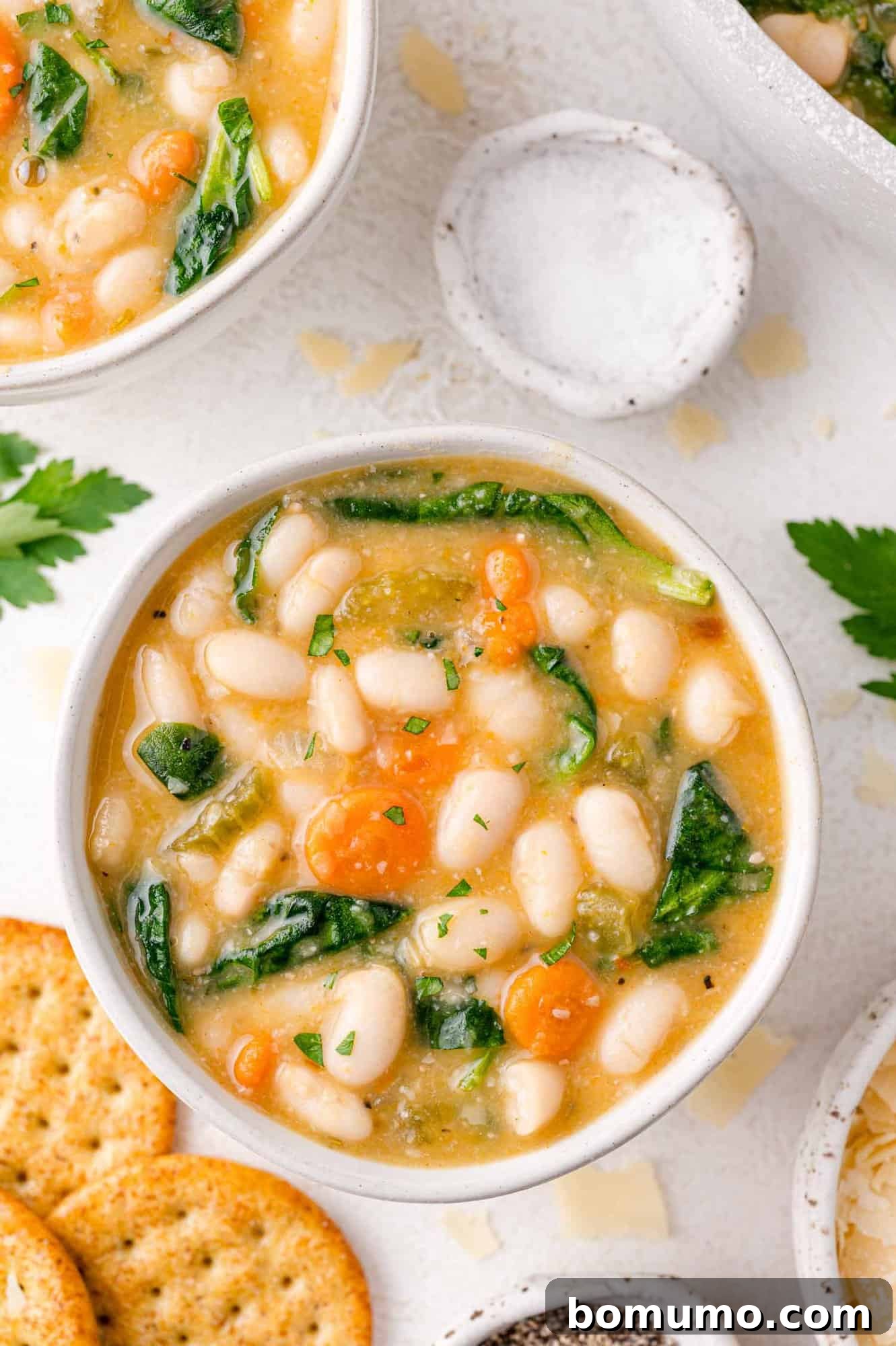 A bowl of white bean and spinach soup with carrots, surrounded by crackers and a small dish of salt.