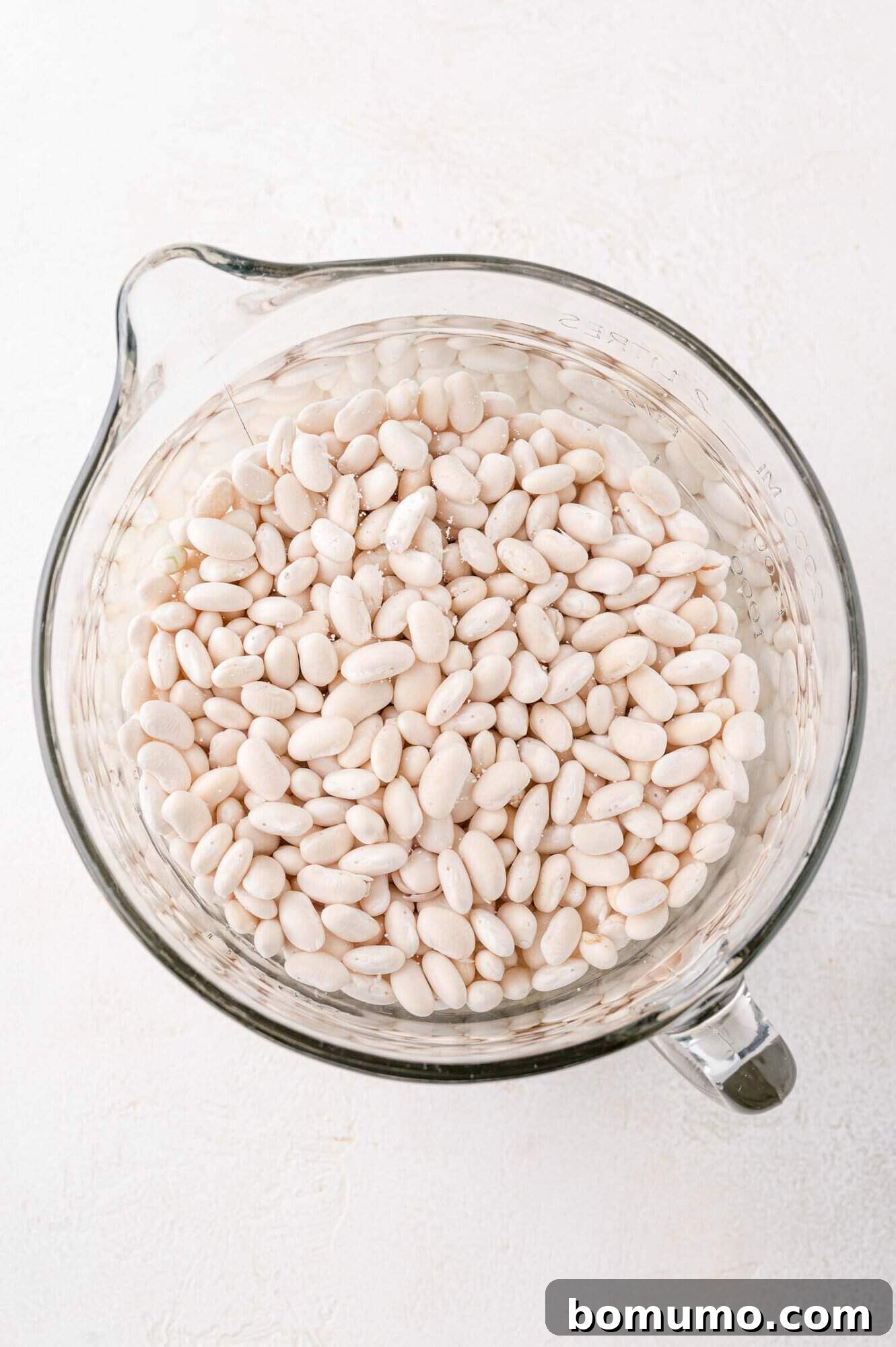 Top view of a glass measuring cup filled with white beans on a white surface.
