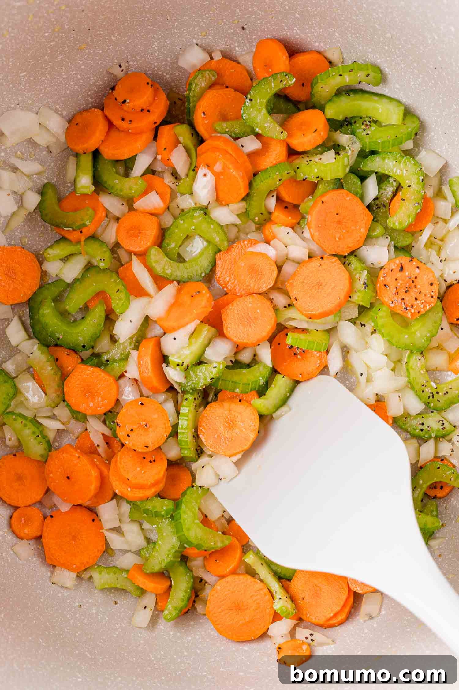 Chopped carrots, celery, and onions sautéed in a pan with ground black pepper, stirred with a white spatula.