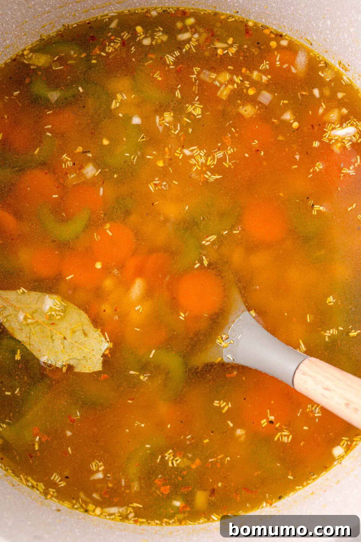 Pot of vegetable soup with carrots, celery, herbs, and a bay leaf, being stirred with a wooden spoon.