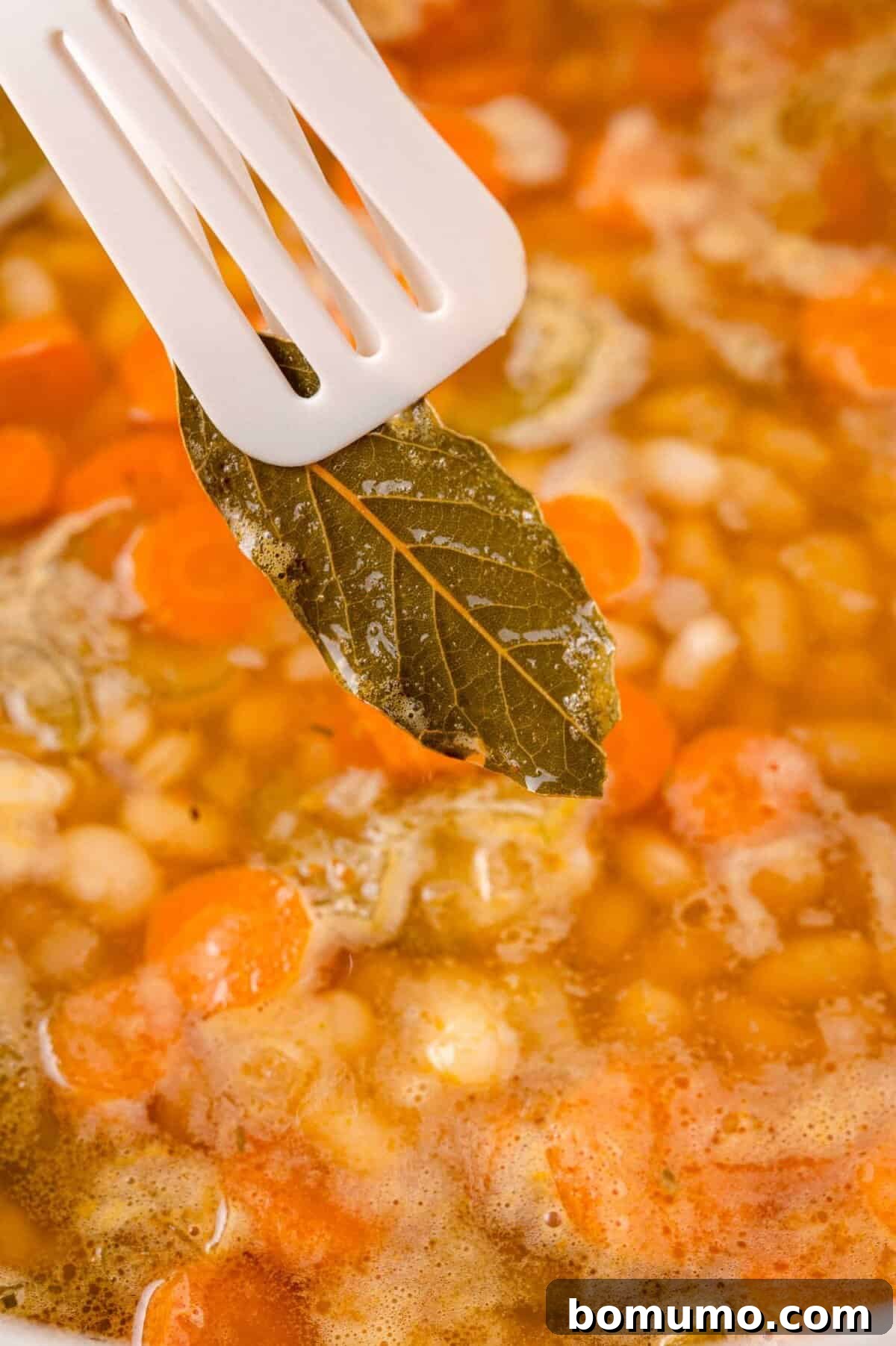 A bay leaf held by a white spatula over a simmering pot of soup with carrots and white beans.