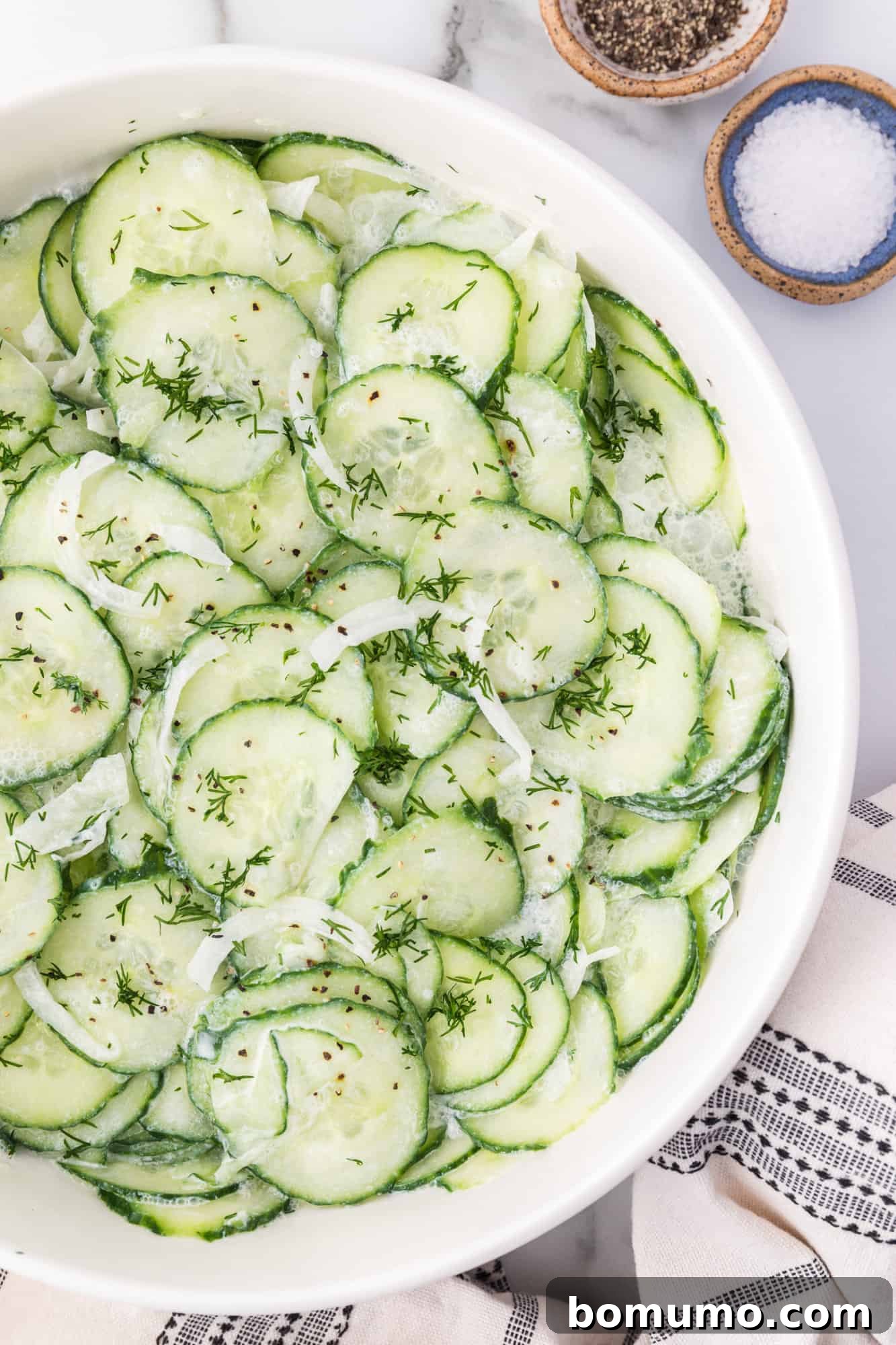 Overhead view of a vibrant German cucumber salad in a pristine white bowl, elegantly arranged beside small ramekins holding salt and pepper, highlighting its fresh appeal.