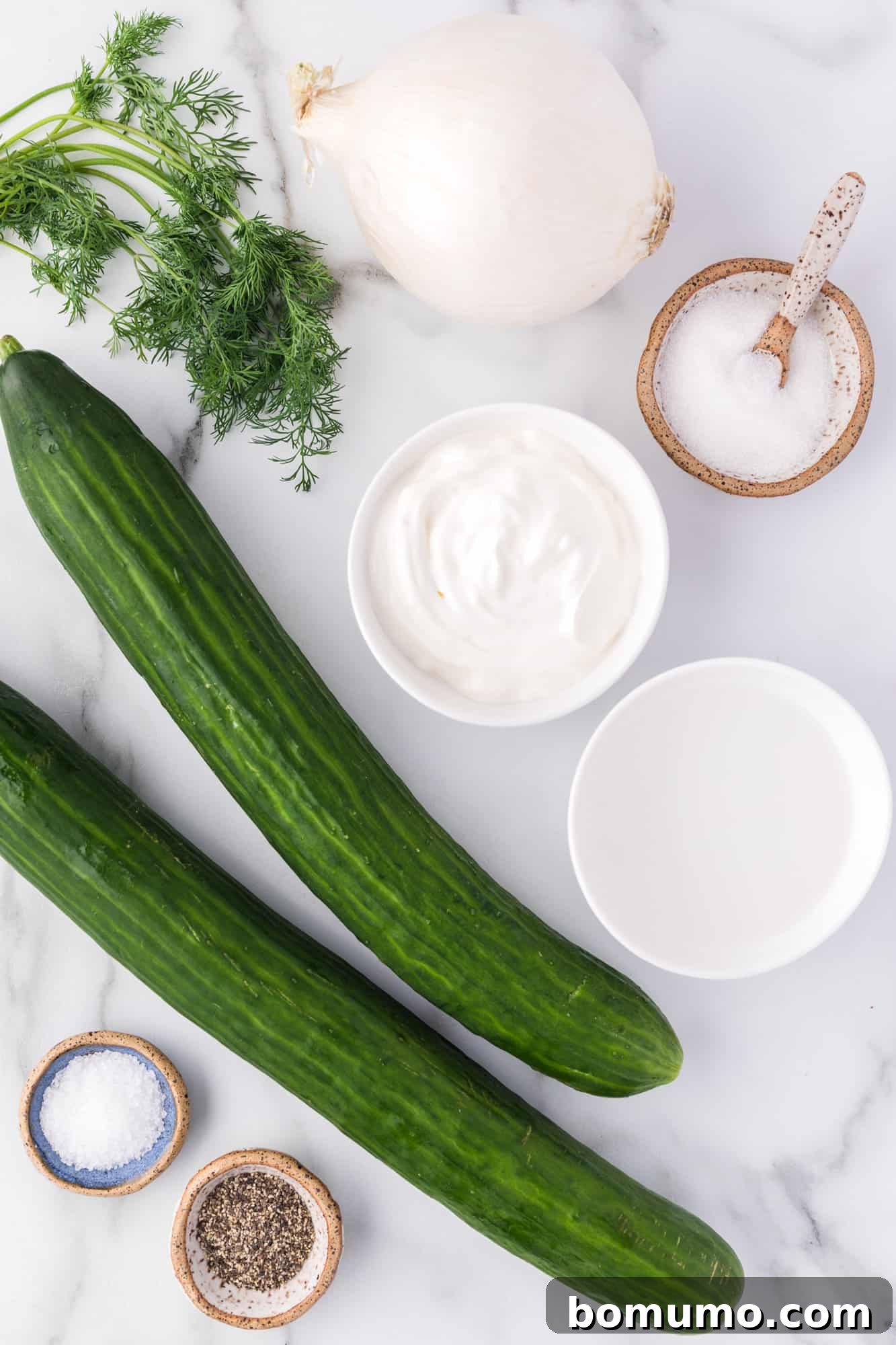 A vibrant display of fresh ingredients for German cucumber salad, featuring crisp cucumbers, a yellow onion, a tub of sour cream, and a bunch of fragrant dill, ready for preparation.