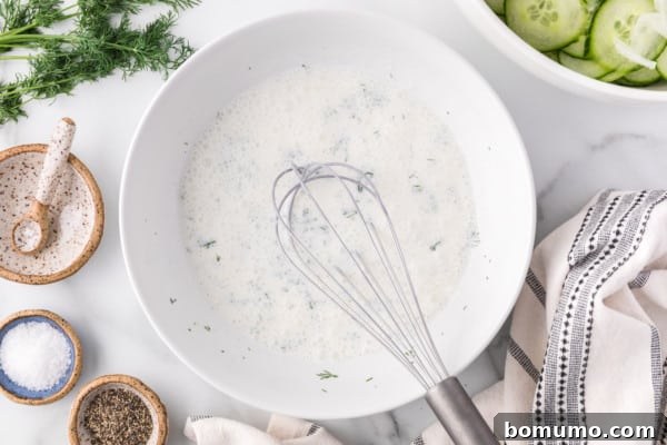 A close-up view of the creamy cucumber salad dressing in a white bowl, freshly whisked to a smooth consistency, with a metal whisk resting inside.