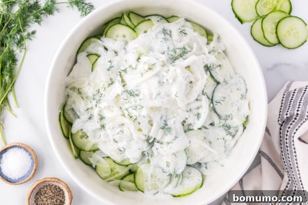A bowl filled with thinly sliced cucumbers, with the creamy dill dressing being generously poured over them, ready for tossing.