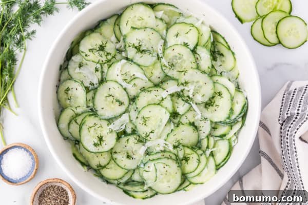 The freshly assembled German cucumber salad in a white bowl, showcasing thinly sliced cucumbers and onions evenly coated in a creamy dill dressing, ready for chilling.