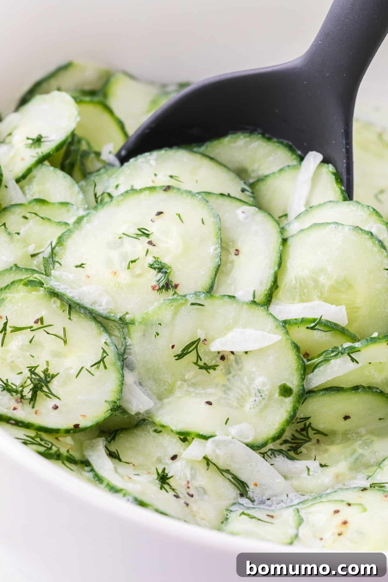 A sleek black serving spoon is used to scoop a portion of fresh German cucumber salad from a large, elegant white bowl, showcasing the creamy texture and vibrant dill.