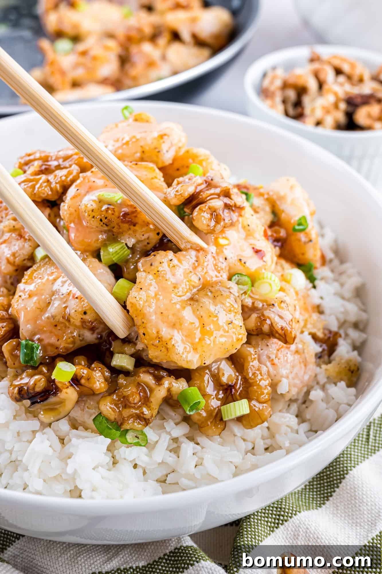 A vibrant bowl of rice topped with golden-brown shrimp and crunchy candied walnuts, artfully garnished with fresh chopped green onions. A pair of elegant chopsticks hovers enticingly above the dish, inviting diners to savor its flavors.
