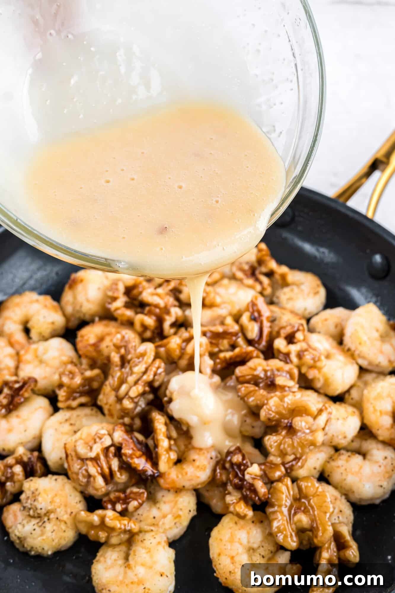 A smooth, creamy sauce being meticulously poured over a skillet filled with freshly cooked shrimp and wholesome walnuts, creating an enticing blend of textures and flavors.